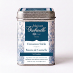 A metal tin of Maison Gabrielle Cinnamon Sticks from Sri Lanka, featuring a blue and white floral pattern with a dark blue label, set against a plain white background.