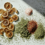 A styled flat-lay of dried citrus slices and piles of ground spices on a white marble surface with dramatic diagonal shadows. The spices include green herbs, a red chili powder pile, and a pink salt pile. MAISON GABRIELLE