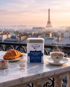 A tin of Maison Gabrielle Petit-Déjeuner Français black tea sits on a marble balcony table with a croissant, a cup of tea, and a Paris cityscape with the Eiffel Tower in the background.