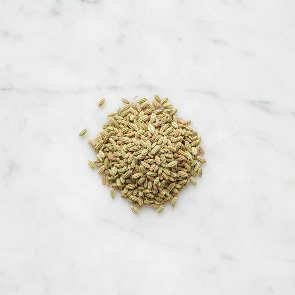 Overhead view of a mound of premium whole fennel seeds from Maison Gabrielle on a bright white marble surface, highlighting the spice's natural green and golden tones and ribbed texture.