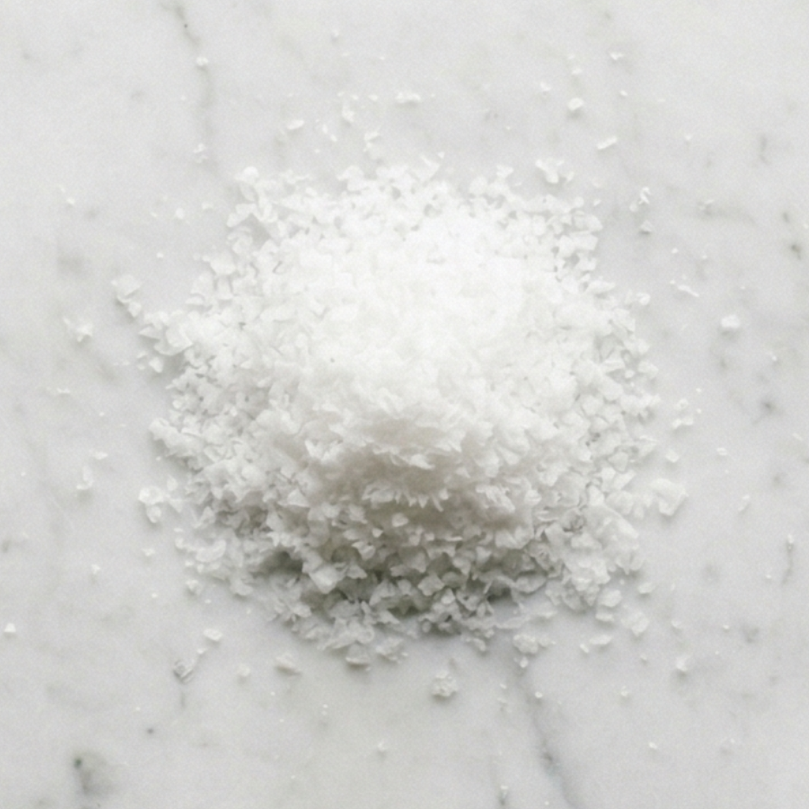 Overhead view of Maison Gabrielle's hand-harvested Fleur de Sel on a white marble surface, showcasing the delicate, crystalline texture and pure white quality of the finishing salt.